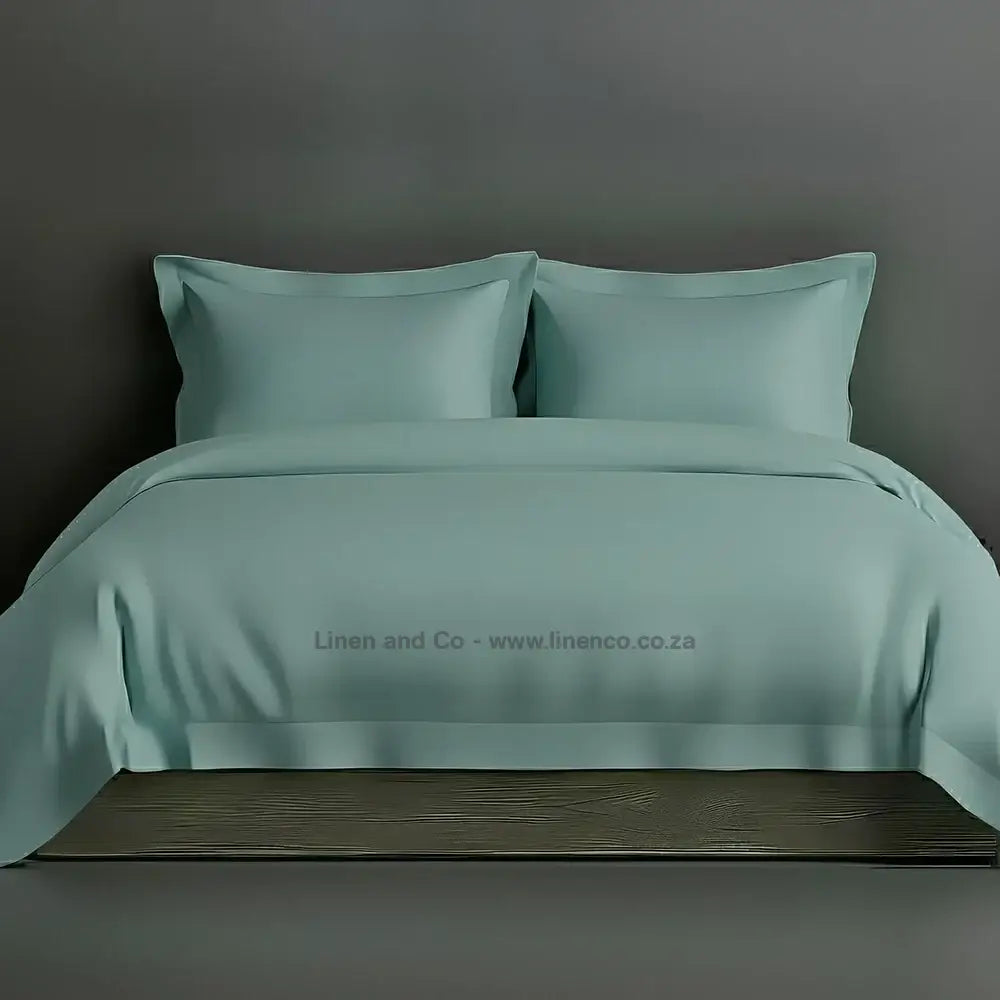 Duck egg percale duvet cover set with Oxford border from Linen and Co, showcasing elegant hospitality bedding with against a dark grey background.