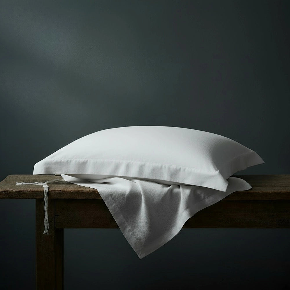 White oxford cotton percale pillowcases stacked on wooden bench against moody dark teal-grey wall—premium plain pillowcases by Linen & Co