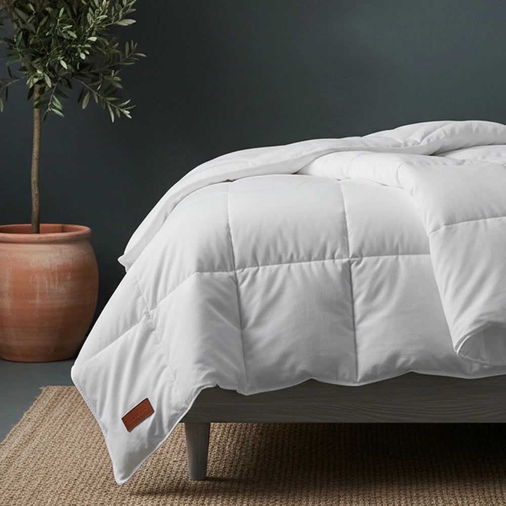 White duvet inner with box-stitch quilting on grey wood bed frame styled with white pillows, olive tree in terracotta pot, and beige jute mat against moody dark teal-grey wall—soft hypoallergenic duvet by Linen & Co