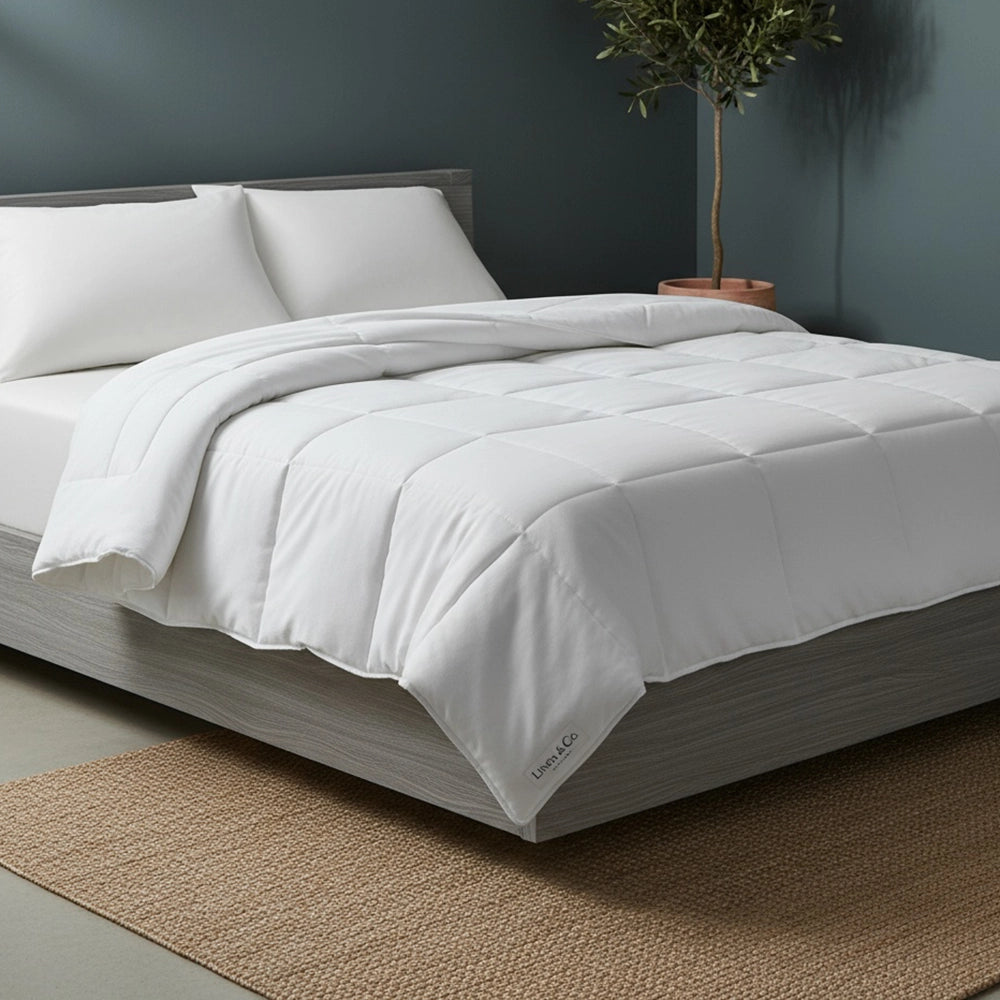 White natural cotton duvet inner on grey wood bed frame styled with white pillows, olive tree in terracotta pot, and beige jute mat against moody dark teal-grey wall—premium breathable bedding by Linen & Co