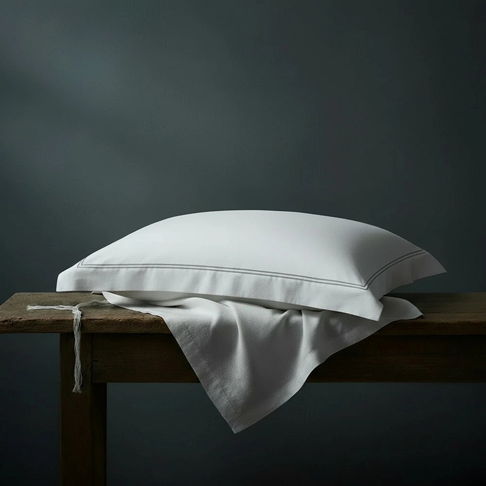 White Oxford pillowcase with grey two-row satin stitch detailing on wooden bench against moody dark teal-grey wall—premium cotton percale pillowcase by Linen & Co