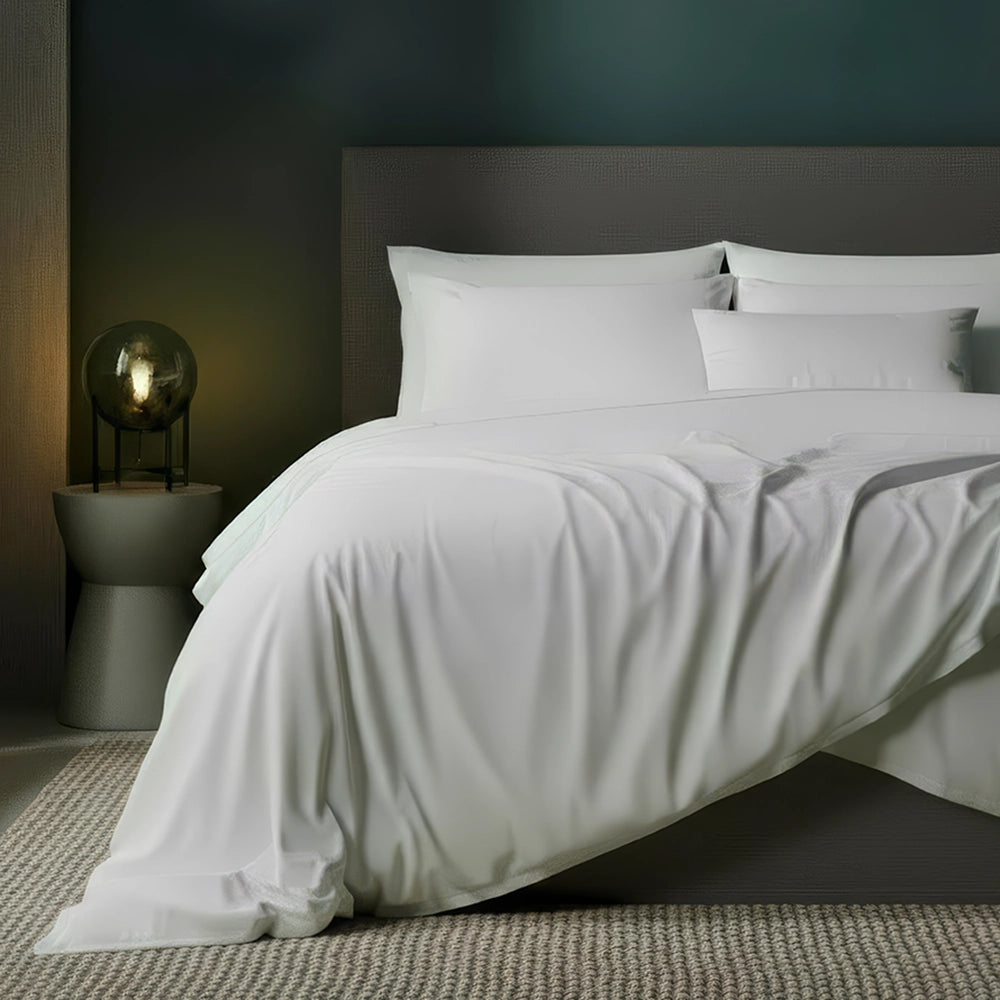 White luxury bedding set on grey bed frame with white pillows and flat sheet, styled with plant and bedside lamp against moody dark teal-grey wall—premium linens by Linen & Co