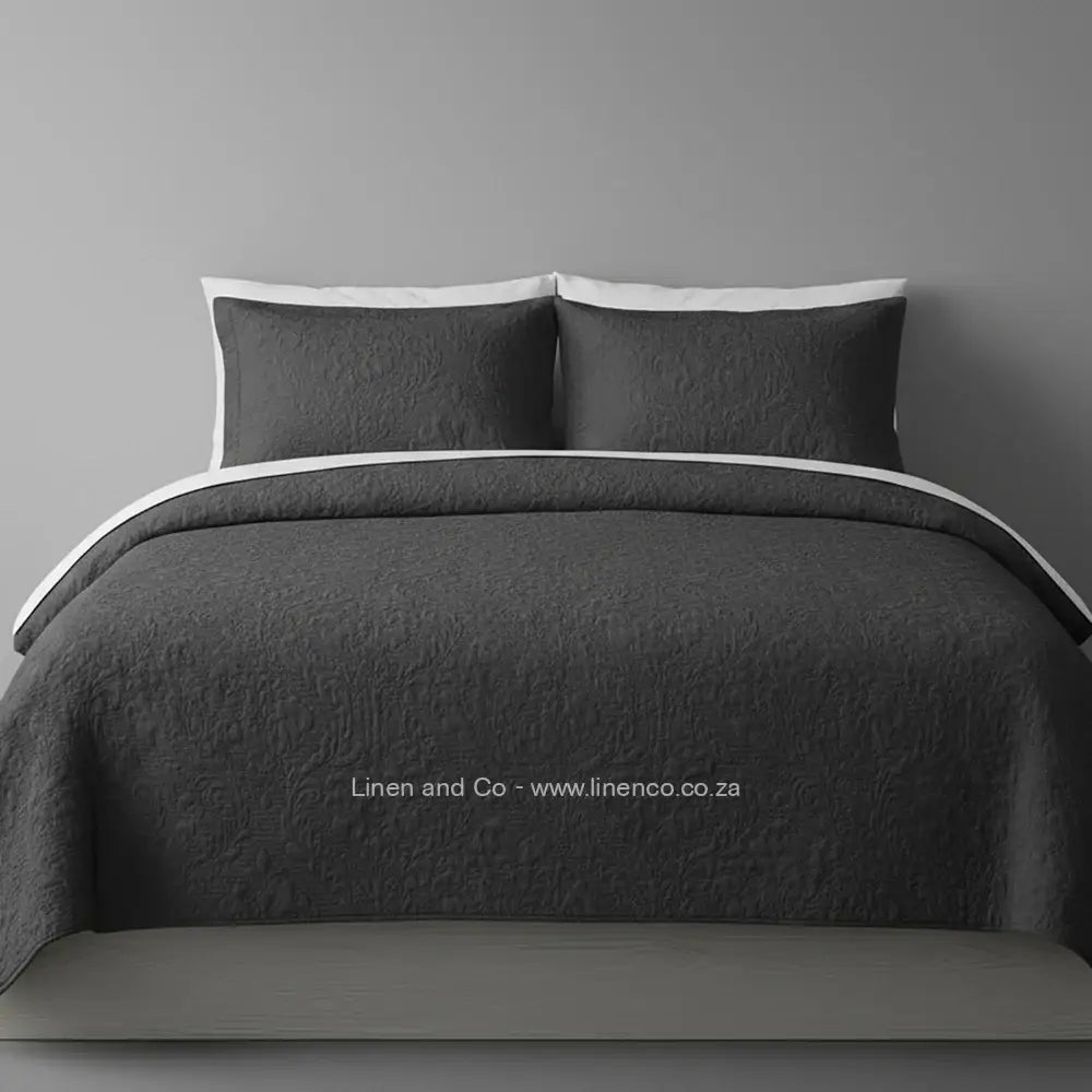 Charcoal Quilted Bedspread Set with Melissa Design, showcasing textured comforter and matching pillow cases on a bed against a dark grey wall, highlighting professional hospitality textile styling by Linen and Co.