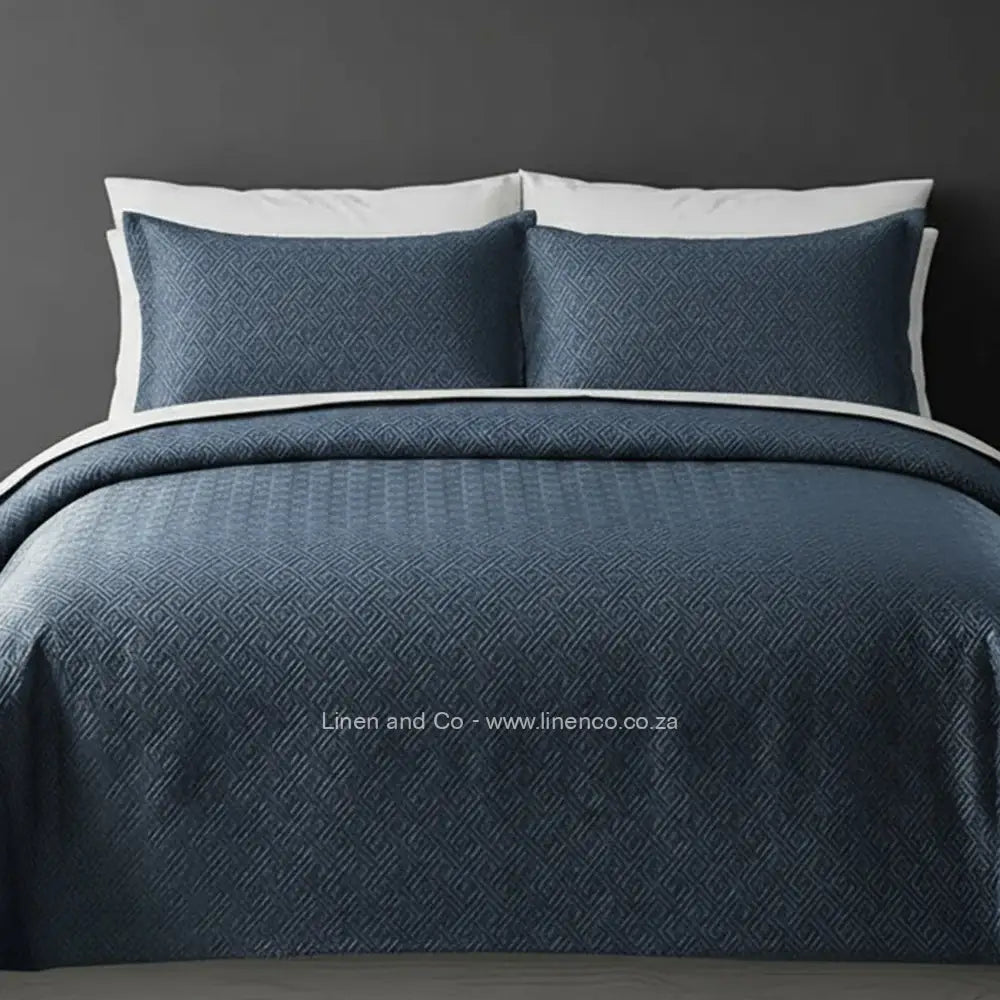 Blue Quilted Bedspread Set with Rome Design, showcasing textured comforter and matching pillow cases on a bed against a dark grey wall, highlighting professional hospitality textile styling by Linen and Co.
