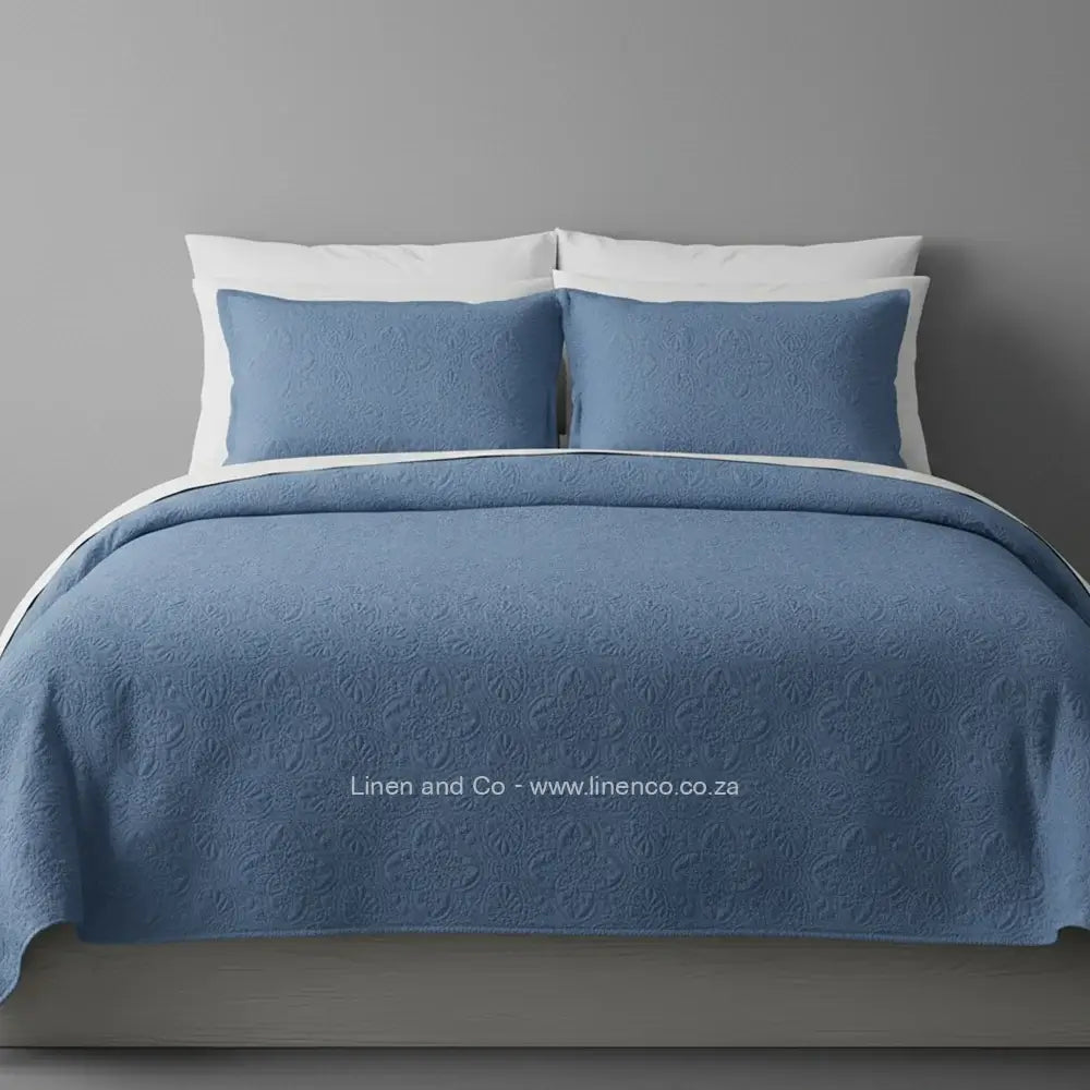 Blue Quilted Bedspread Set with Daisy Design, showcasing textured comforter and matching pillow cases on a bed against a dark grey wall, highlighting professional hospitality textile styling by Linen and Co.