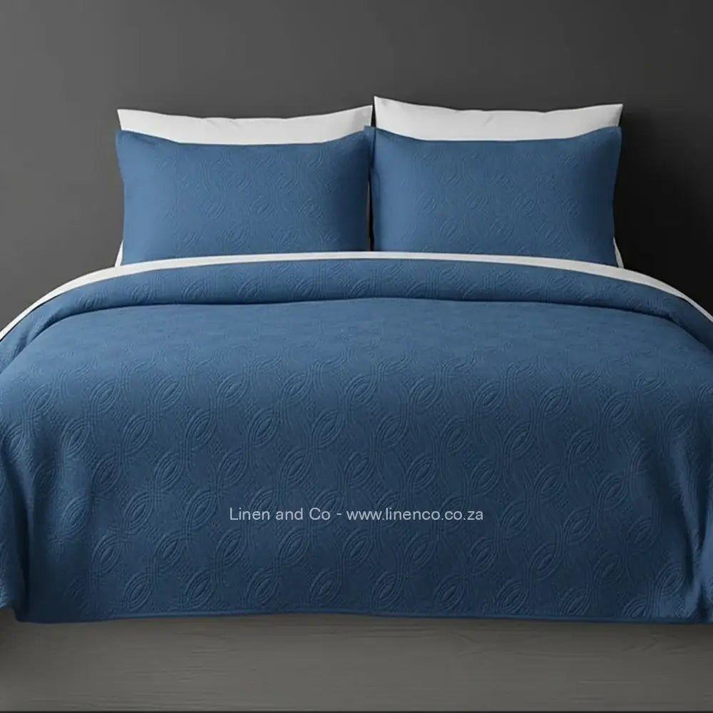 Blue Quilted Bedspread Set with Munich Design, showcasing textured comforter and matching pillow cases on a bed against a dark grey wall, highlighting professional hospitality textile styling by Linen and Co.