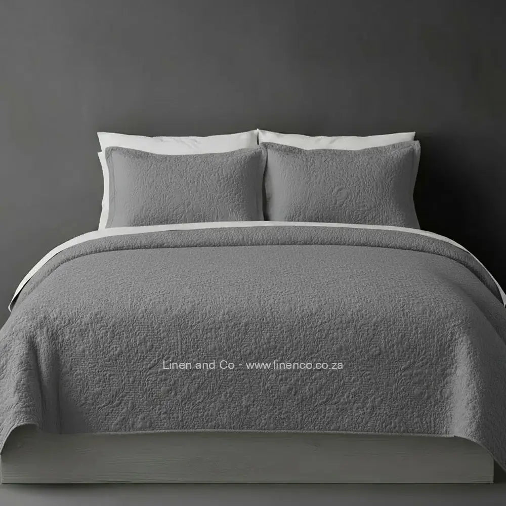Grey Quilted Bedspread Set with Carmen Design, showcasing textured comforter and matching pillow cases on a bed against a dark grey wall, highlighting professional hospitality textile styling by Linen and Co.