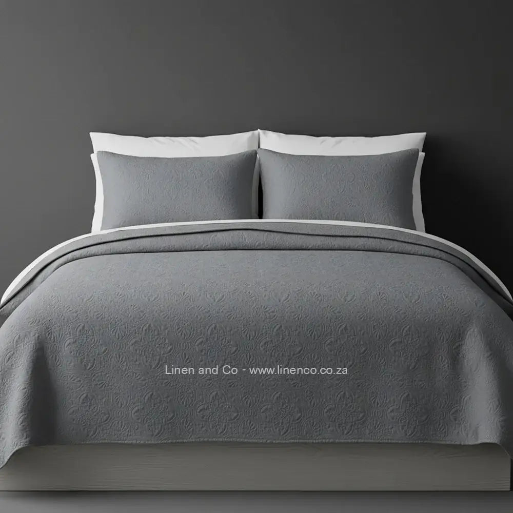 Grey Quilted Bedspread Set with Daisy Design, showcasing textured comforter and matching pillow cases on a bed against a dark grey wall, highlighting professional hospitality textile styling by Linen and Co.