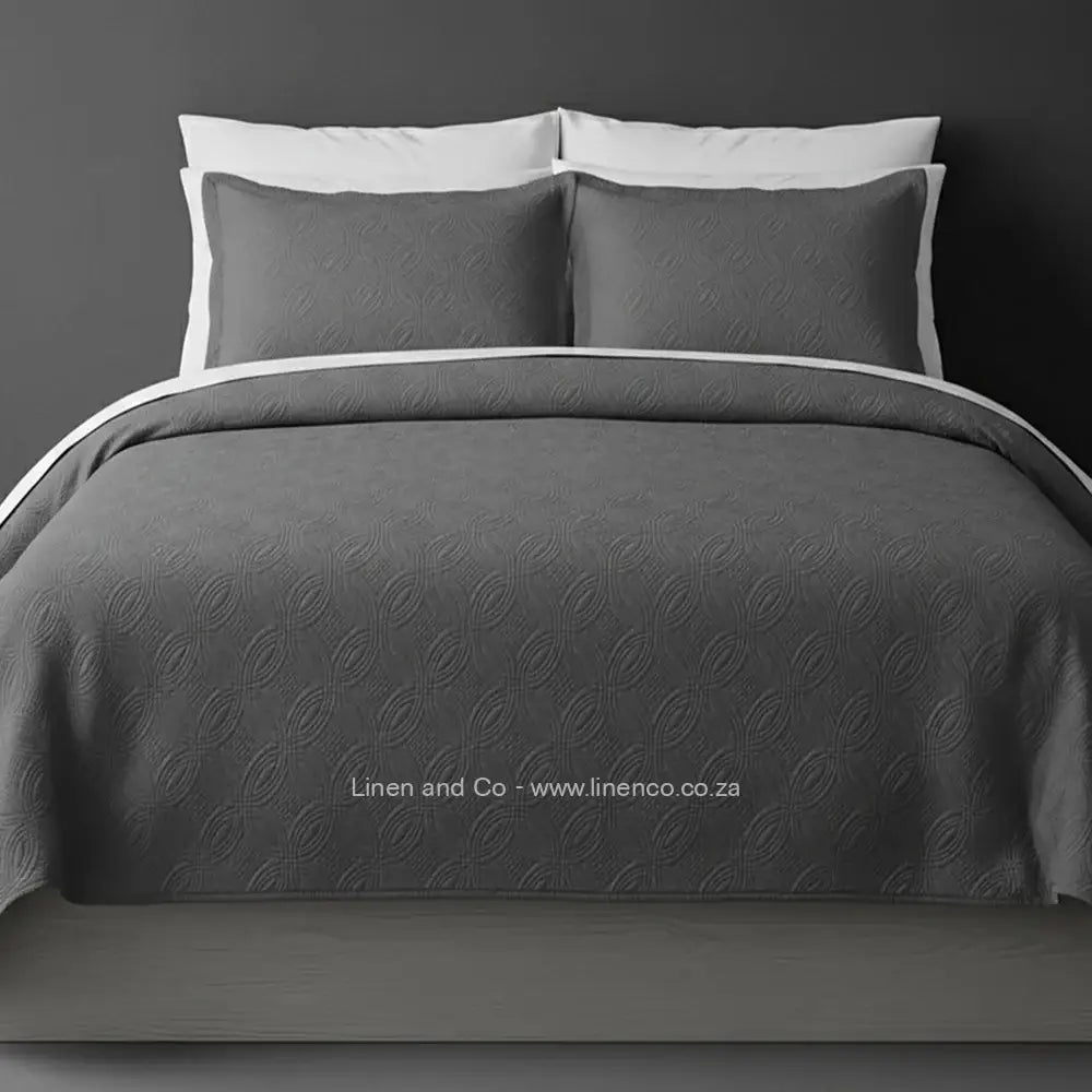 Grey Quilted Bedspread Set with Munich Design, showcasing textured comforter and matching pillow cases on a bed against a dark grey wall, highlighting professional hospitality textile styling by Linen and Co.