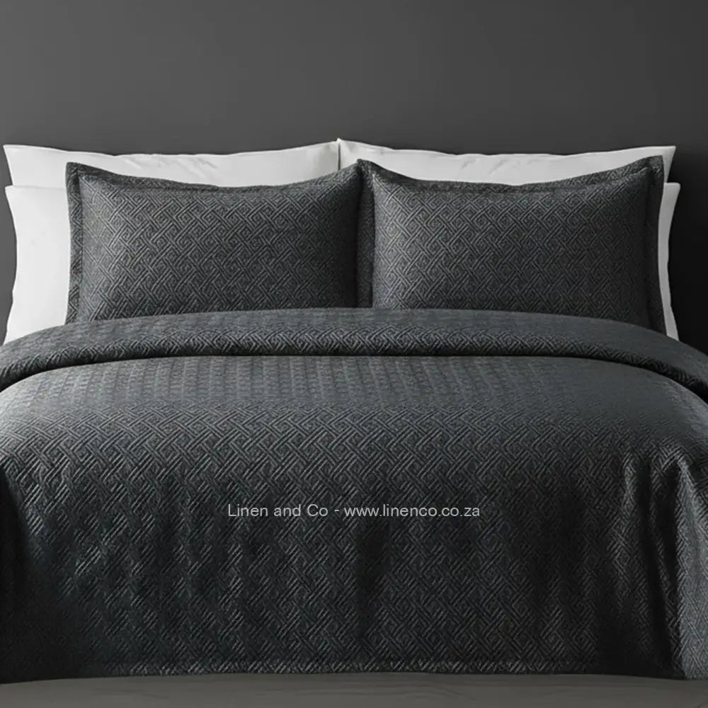 Dark Grey Quilted Bedspread Set with Rome Design, showcasing textured comforter and matching pillow cases on a bed against a dark grey wall, highlighting professional hospitality textile styling by Linen and Co.