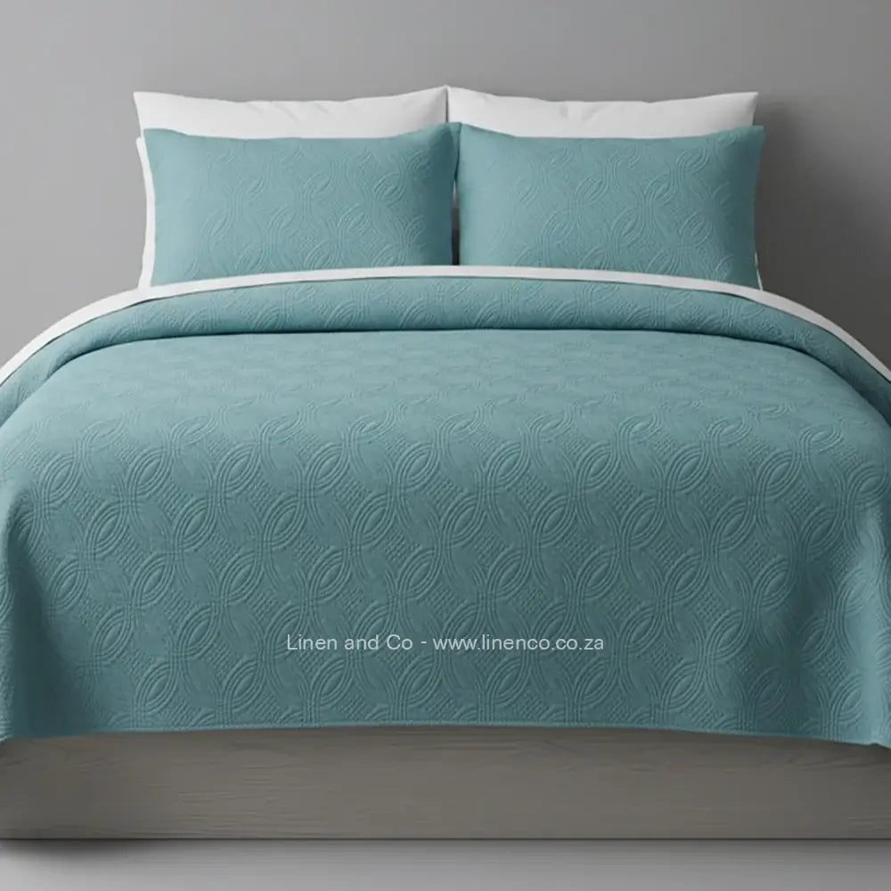 Teal Quilted Bedspread Set with Munich Design, showcasing textured comforter and matching pillow cases on a bed against a dark grey wall, highlighting professional hospitality textile styling by Linen and Co.