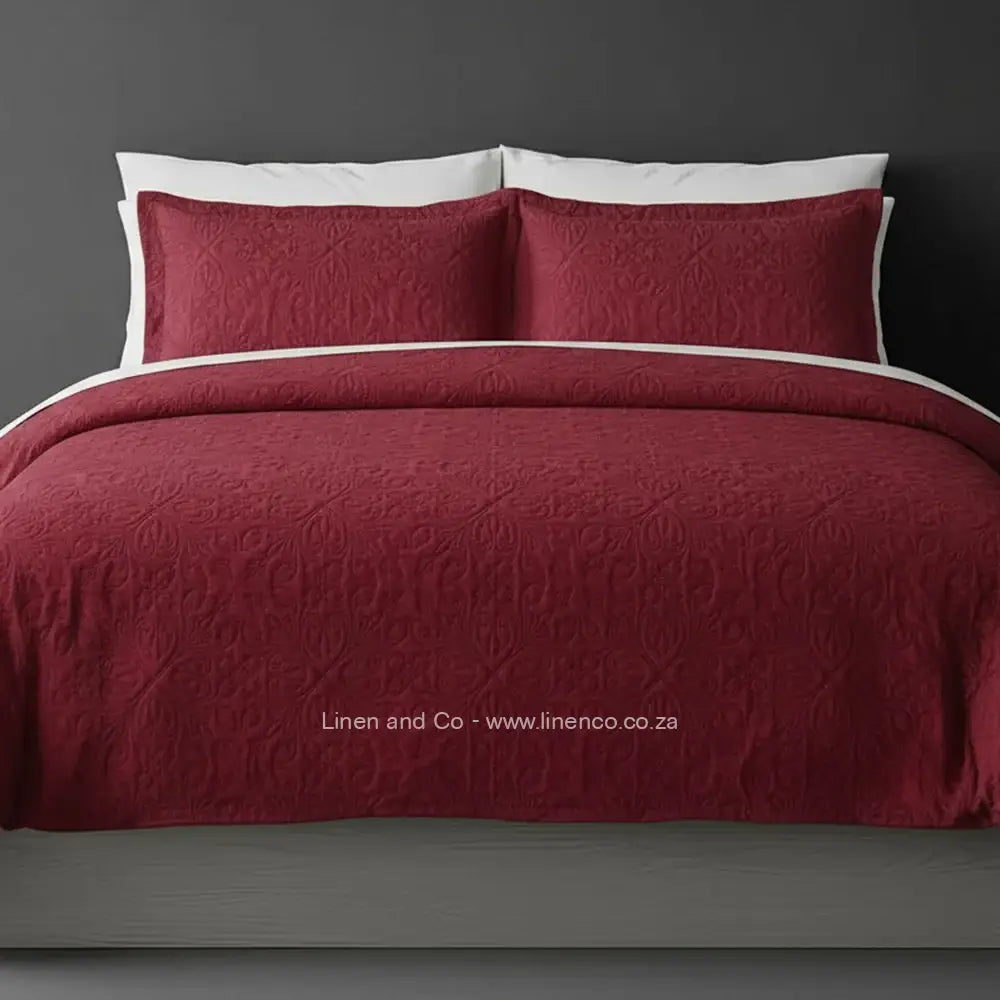 Red/maroon Quilted Bedspread Set with French Velvet, showcasing textured comforter and matching pillow cases on a bed against a dark grey wall, highlighting professional hospitality textile styling by Linen and Co.