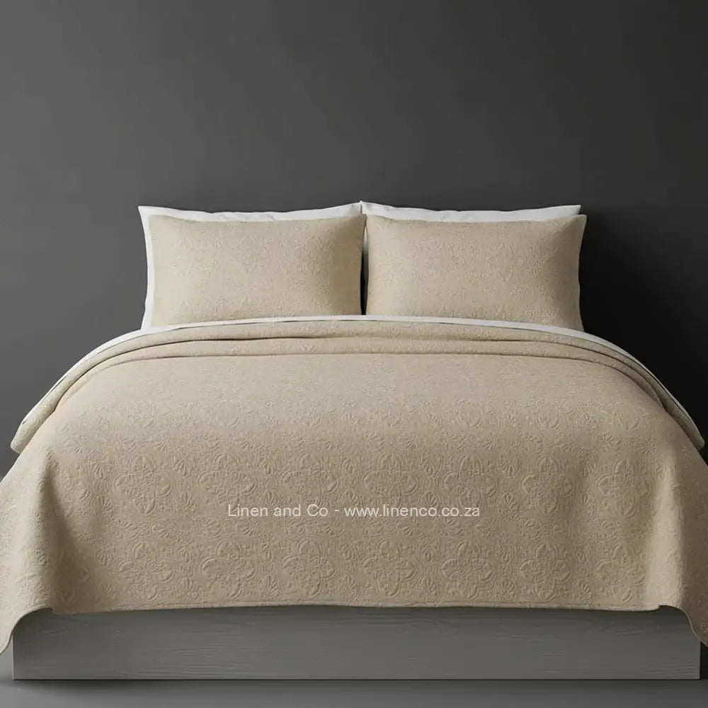 Beige Quilted Bedspread Set with Daisy Design, showcasing textured comforter and matching pillow cases on a bed against a dark grey wall, highlighting professional hospitality textile styling by Linen and Co.