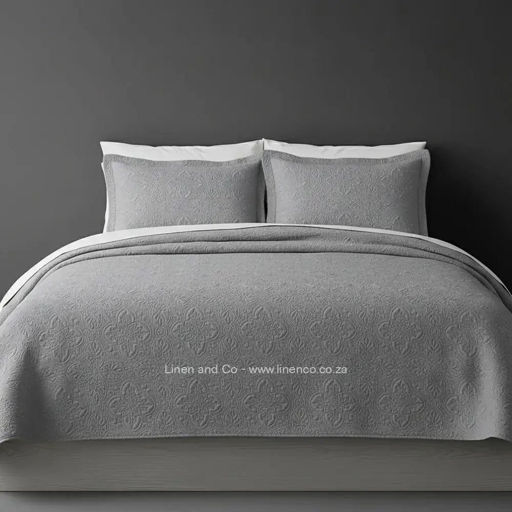 Grey Quilted Bedspread Set with Daisy Design, showcasing textured comforter and matching pillow cases on a bed against a dark grey wall, highlighting professional hospitality textile styling by Linen and Co.