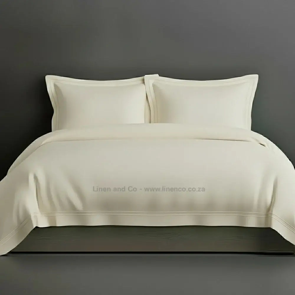 Cream percale duvet cover set with Oxford two-row white satin stitch border from Linen and Co, showcasing elegant hospitality bedding with subtle decorative detailing against a dark grey background.