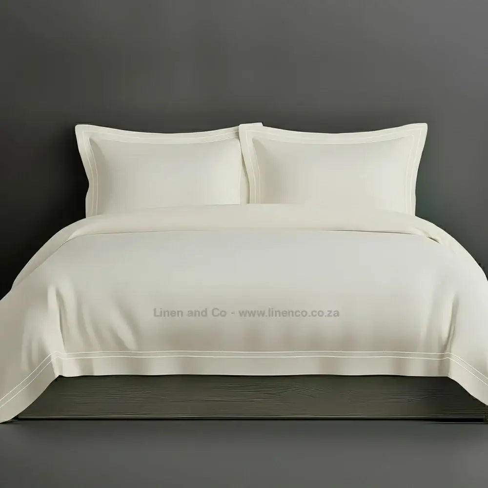 Cream percale duvet cover set with Oxford two-row white satin stitch border from Linen and Co, showcasing elegant hospitality bedding with subtle decorative detailing against a dark grey background.