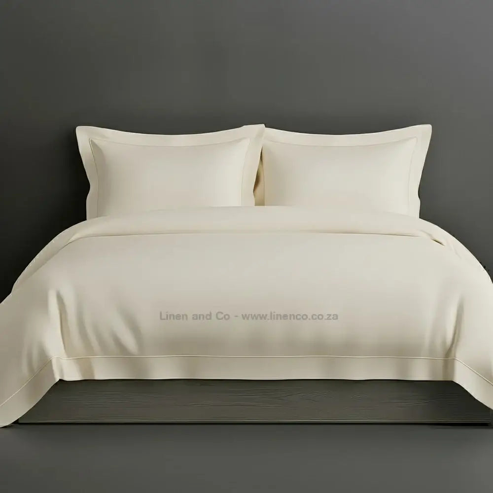 Cream percale duvet cover set with Oxford two-row white satin stitch border from Linen and Co, showcasing elegant hospitality bedding with subtle decorative detailing against a dark grey background.