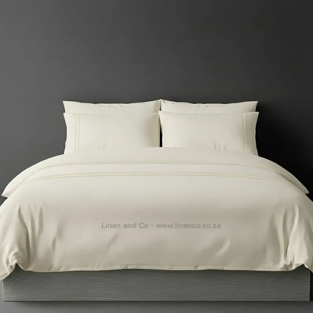 Cream polycotton percale duvet cover set with two-row white satin stitch border from Linen and Co, showcasing elegant hospitality bedding with subtle decorative detailing against a dark grey background.