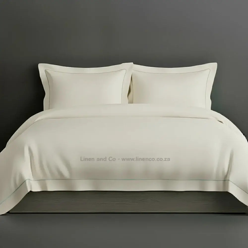 Cream percale duvet cover set with Oxford two-row white satin stitch border from Linen and Co, showcasing elegant hospitality bedding with subtle decorative detailing against a dark grey background.