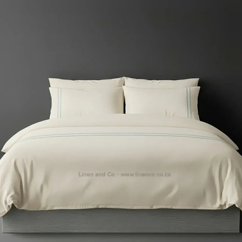 Cream polycotton percale duvet cover set with two-row white satin stitch border from Linen and Co, showcasing elegant hospitality bedding with subtle decorative detailing against a dark grey background