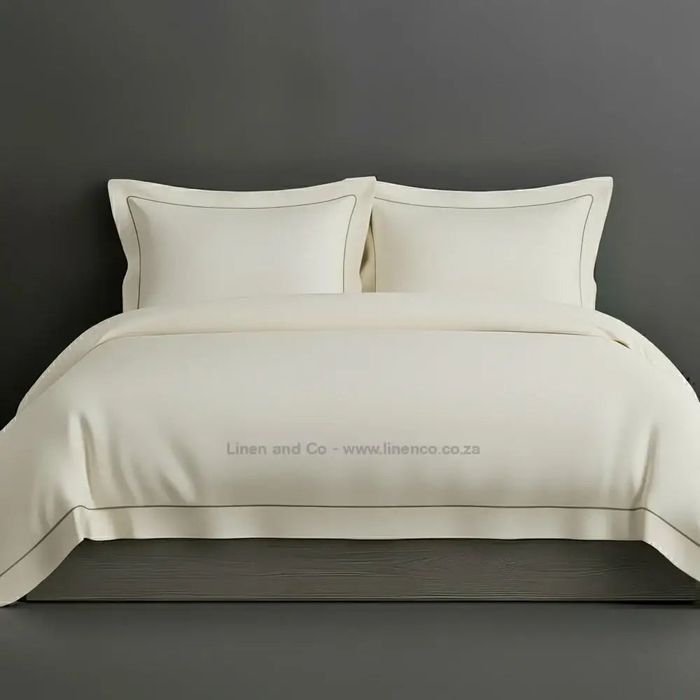 Cream percale duvet cover set with Oxford two-row white satin stitch border from Linen and Co, showcasing elegant hospitality bedding with subtle decorative detailing against a dark grey background.