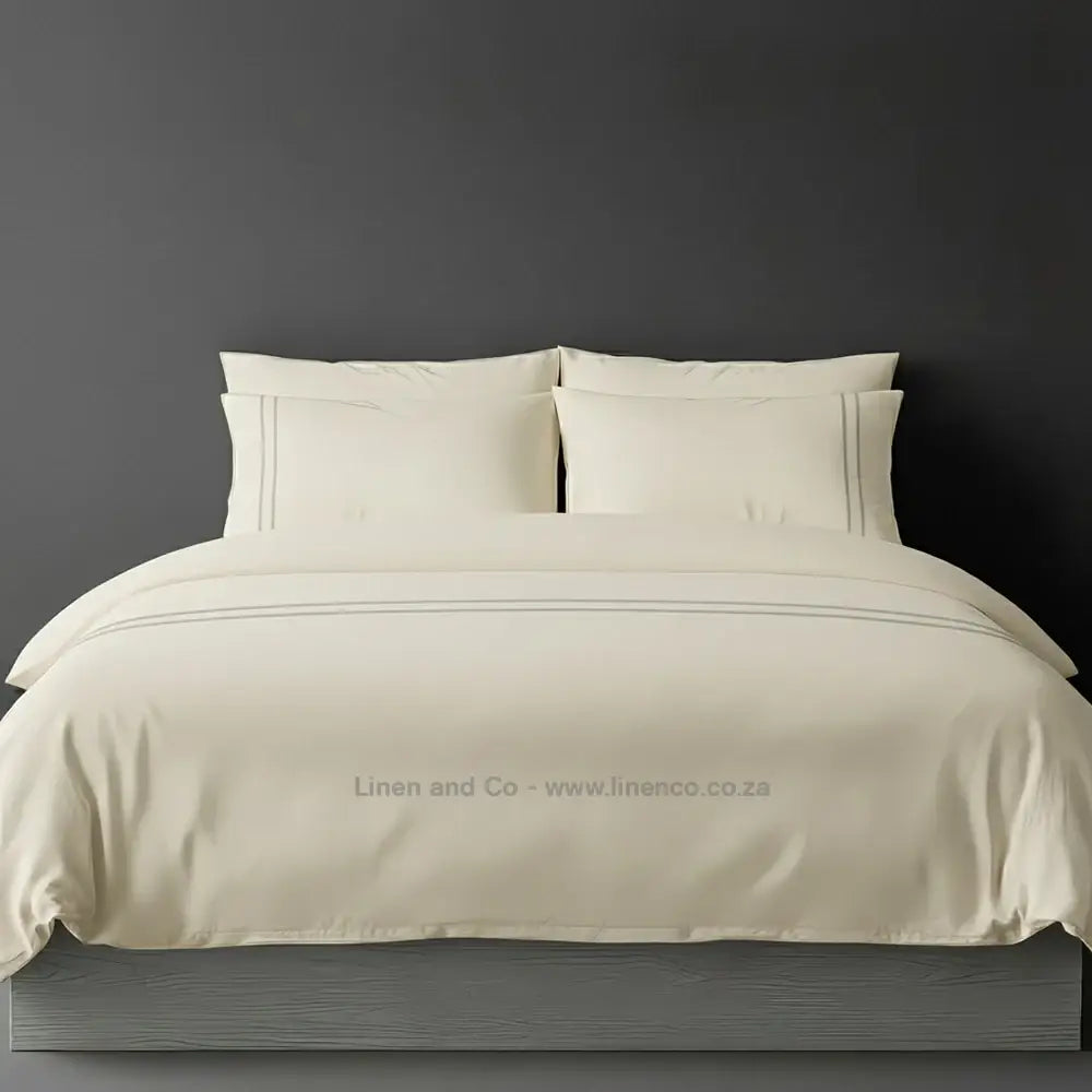 Cream percale duvet cover set with two-row white satin stitch border from Linen and Co, showcasing elegant hospitality bedding with subtle decorative detailing against a dark grey background