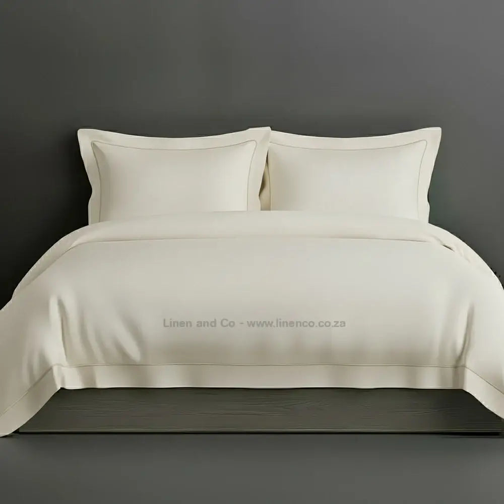 Cream percale duvet cover set with Oxford two-row white satin stitch border from Linen and Co, showcasing elegant hospitality bedding with subtle decorative detailing against a dark grey background.