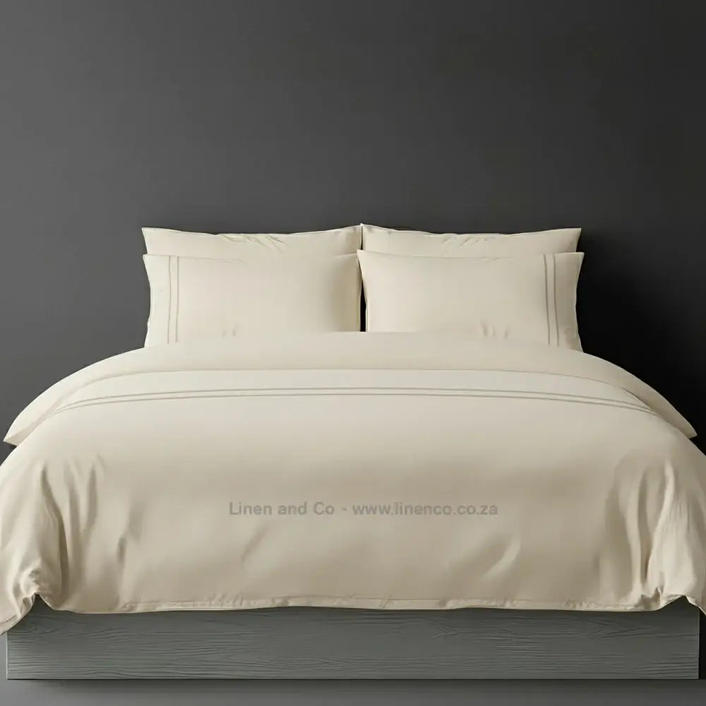 Cream percale duvet cover set with two-row white satin stitch border from Linen and Co, showcasing elegant hospitality bedding with subtle decorative detailing against a dark grey background
