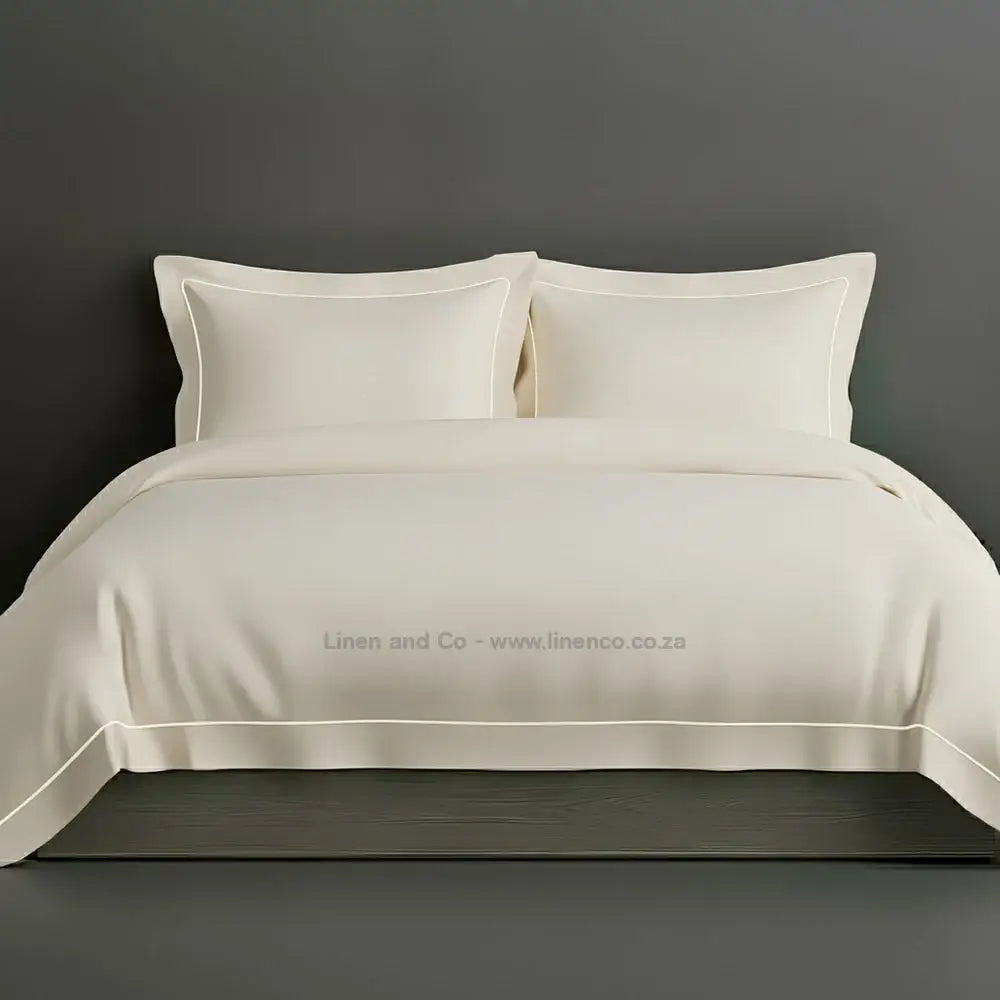 Cream percale duvet cover set with Oxford two-row white satin stitch border from Linen and Co, showcasing elegant hospitality bedding with subtle decorative detailing against a dark grey background.