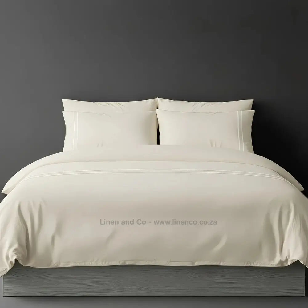 Cream percale duvet cover set with two-row white satin stitch border from Linen and Co, showcasing elegant hospitality bedding with subtle decorative detailing against a dark grey background