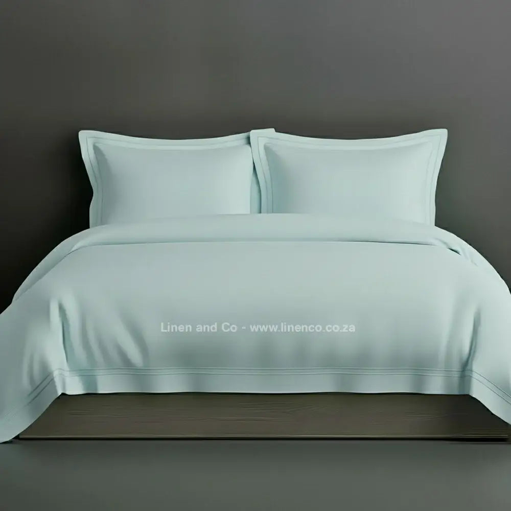 Duck egg percale duvet cover set with Oxford two-row white satin stitch border from Linen and Co, showcasing elegant hospitality bedding with subtle decorative detailing against a dark grey background.