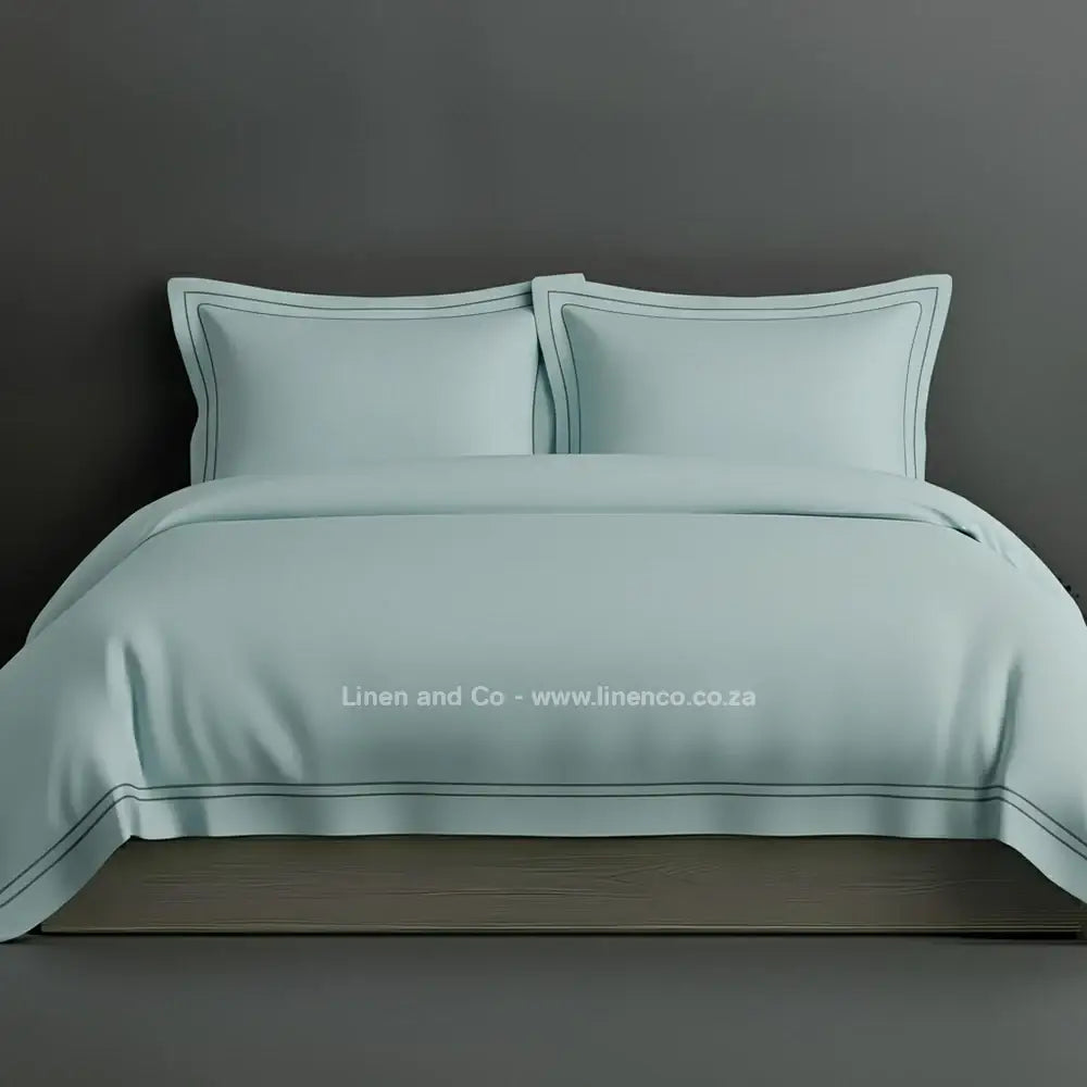 Duck egg percale duvet cover set with Oxford two-row white satin stitch border from Linen and Co, showcasing elegant hospitality bedding with subtle decorative detailing against a dark grey background.