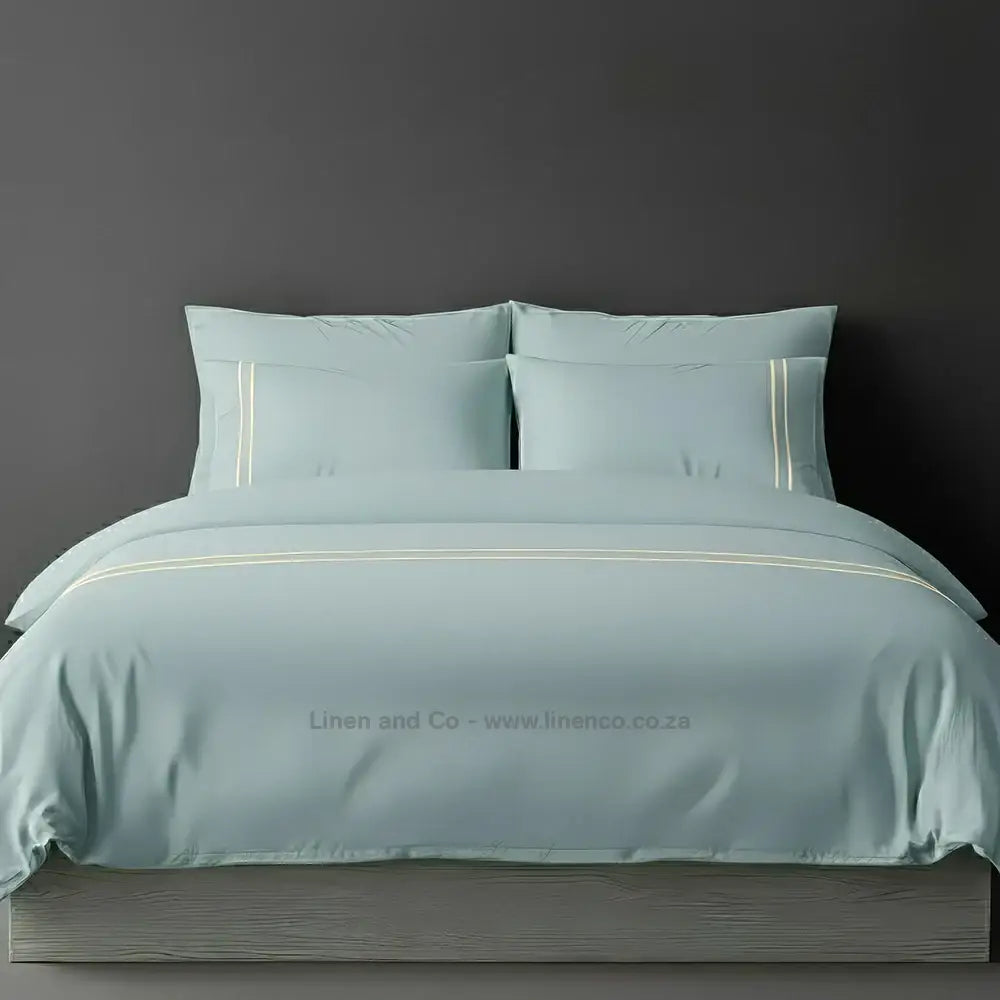 Duck Egg percale duvet cover set with two-row white satin stitch border from Linen and Co, showcasing elegant hospitality bedding with subtle decorative detailing against a dark grey background.