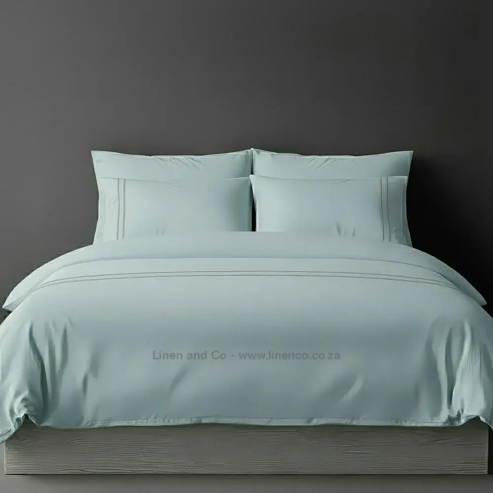 Duck Egg percale duvet cover set with two-row white satin stitch border from Linen and Co, showcasing elegant hospitality bedding with subtle decorative detailing against a dark grey background.