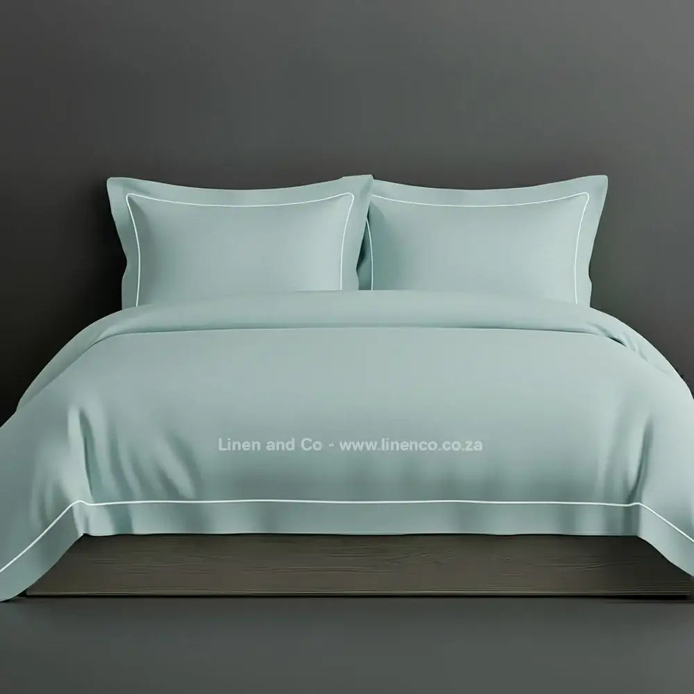 Duck Egg percale duvet cover set with Oxford one-row white satin stitch border from Linen and Co, showcasing elegant hospitality bedding with subtle decorative detailing against a dark grey background.