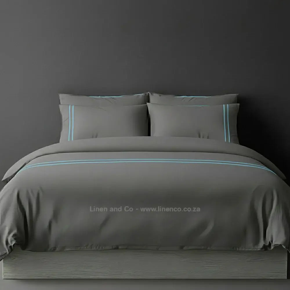 Grey percale duvet cover set with two-row white satin stitch border from Linen and Co, showcasing elegant hospitality bedding with subtle decorative detailing against a dark grey background.