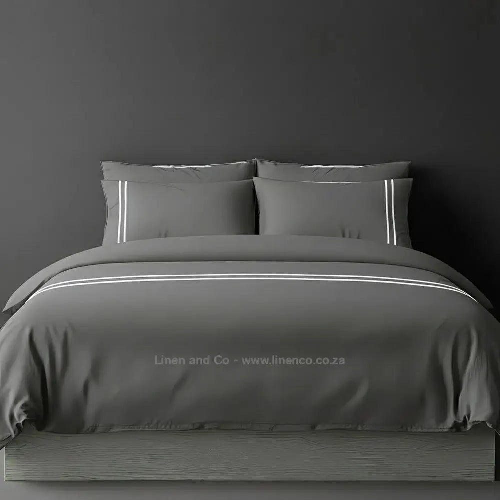 Grey percale duvet cover set with two-row white satin stitch border from Linen and Co, showcasing elegant hospitality bedding with subtle decorative detailing against a dark grey background.