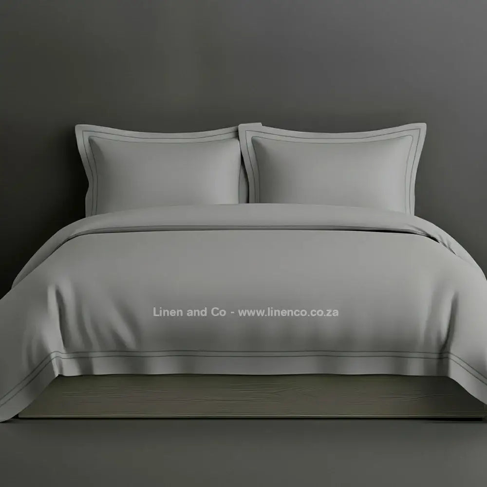 Grey percale duvet cover set with Oxford two-row white satin stitch border from Linen and Co, showcasing elegant hospitality bedding with subtle decorative detailing against a dark grey background.