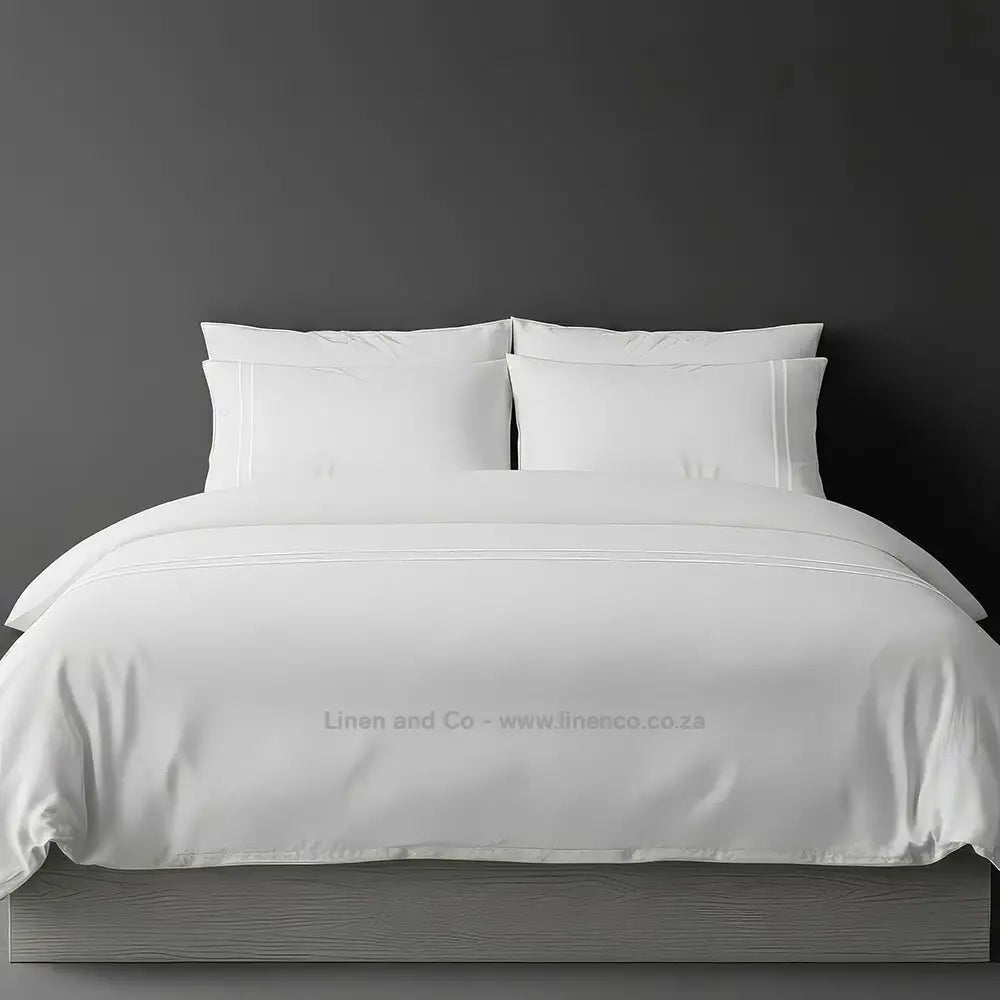 White polycotton percale duvet cover set with two-row white satin stitch border from Linen and Co, showcasing elegant hospitality bedding with subtle decorative detailing against a dark grey background
