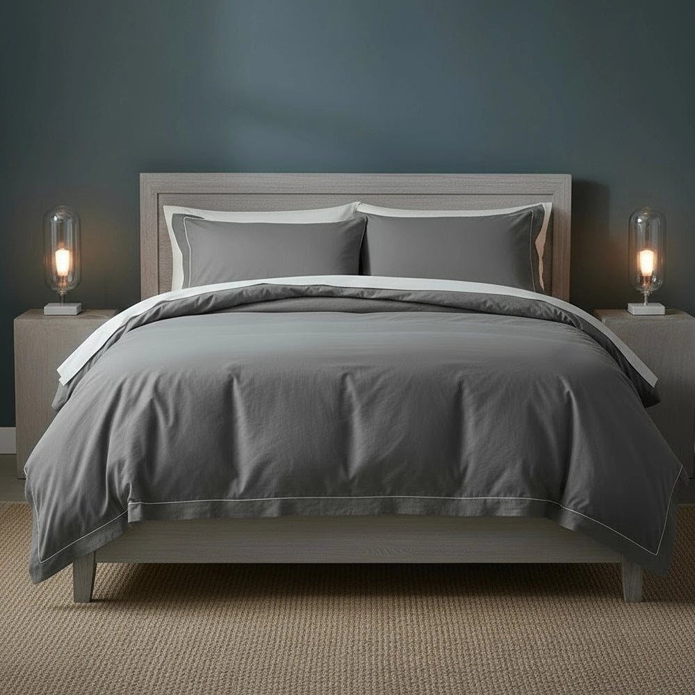 Grey duvet cover with white one-row satin stitch detailing on light grey upholstered bed frame with matching pillowcases, styled with bedside lamps and beige jute mat against moody dark teal-grey wall—premium cotton bedding by Linen & Co