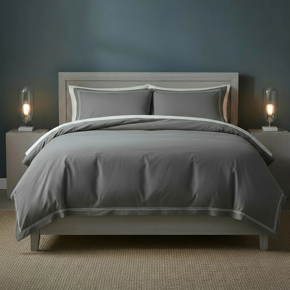Grey duvet cover with white two-row satin stitch detailing on light grey upholstered bed frame with matching pillowcases, styled with bedside lamps and beige jute mat against moody dark teal-grey wall—premium cotton bedding by Linen & Co