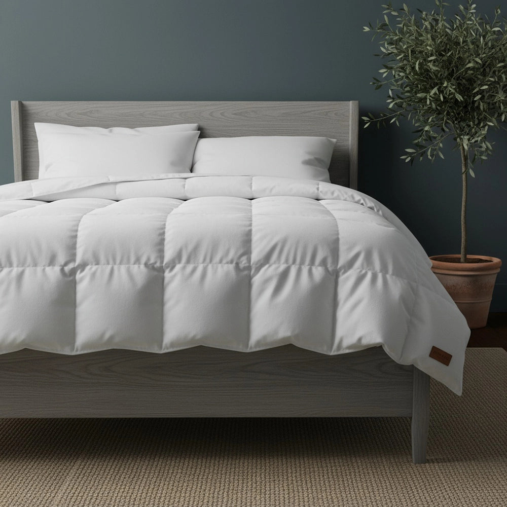 White goose down duvet inner with box-stitch quilting on grey wood bed frame styled with white pillows, olive tree in terracotta pot, and beige jute mat against moody dark teal-grey wall—premium luxury duvet by Linen & Co