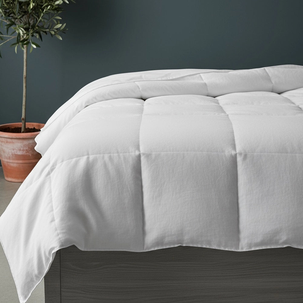 White duvet inner with box-stitch quilting on grey wood bed frame styled with white pillows, olive tree in terracotta pot, and beige jute mat against moody dark teal-grey wall—soft hypoallergenic duvet by Linen & Co