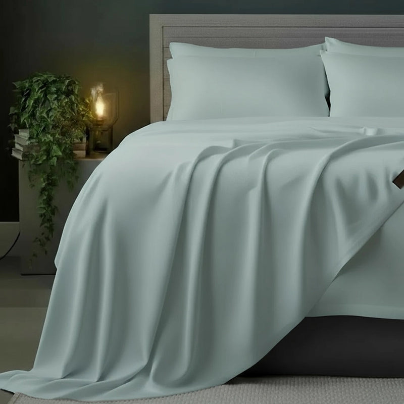 Duck egg flat sheet draped over grey bed frame with white pillows, styled with olive plant and bedside lamp against moody dark teal-grey wall—premium cotton percale flat sheet by Linen & Co