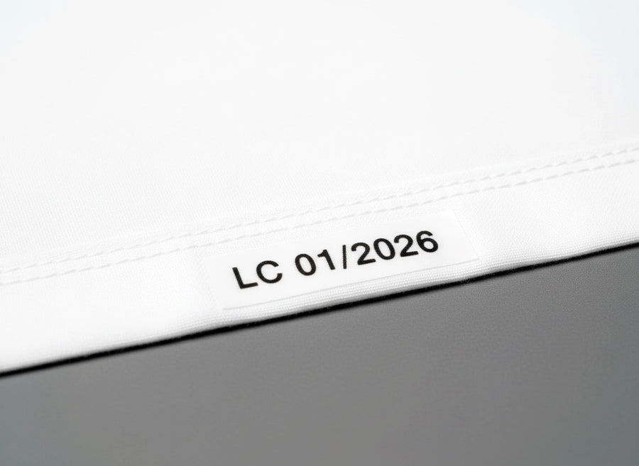 Premium white cotton bedding folded to show inside bottom hem with heat transfer printed text displaying abbreviated property name "LC" and date "01/2026" in black showing utility marking detail