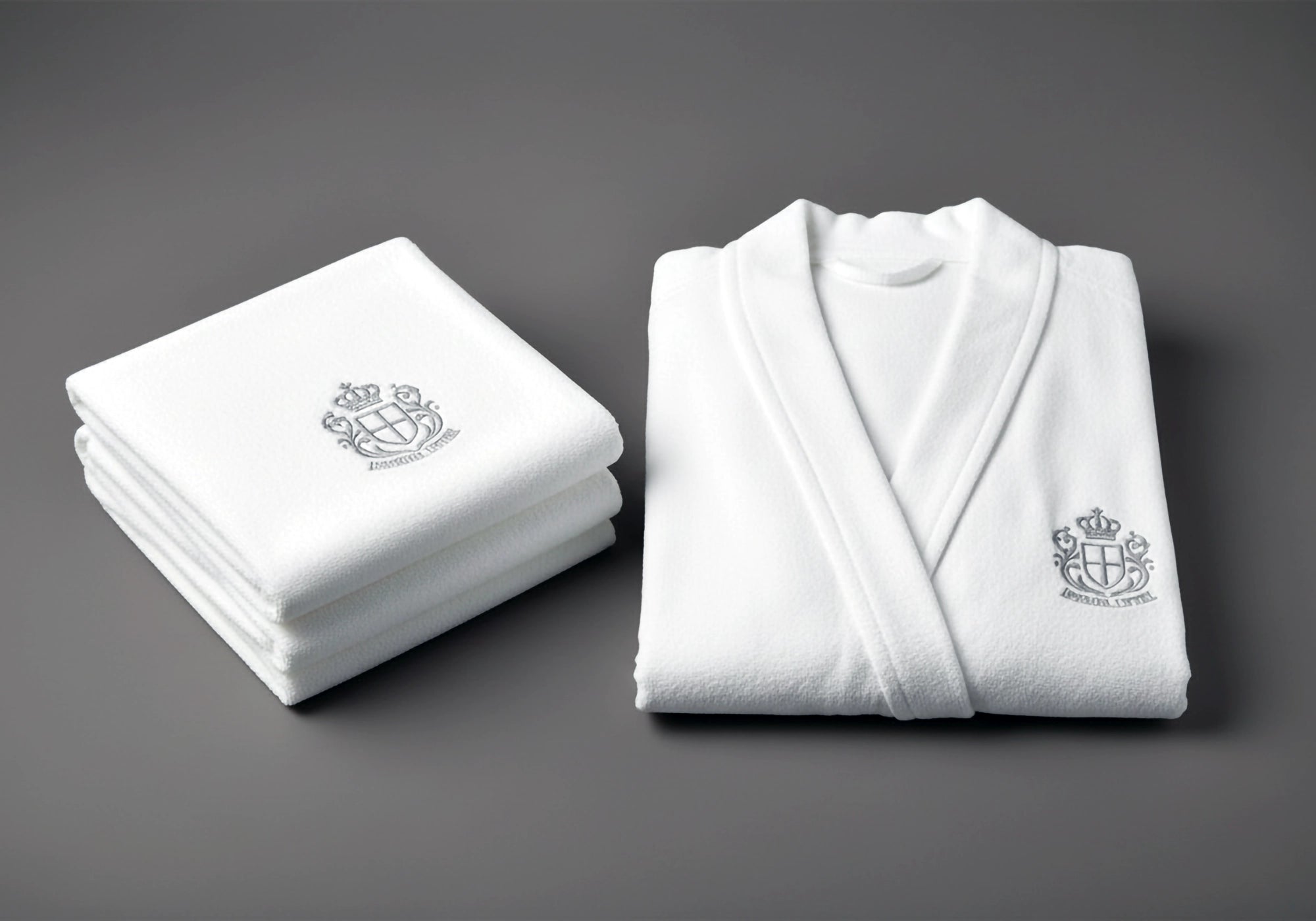 White embroidered bathrobe and folded towels on a grey background by Linen and Co - premium hospitality bath linen with custom branding