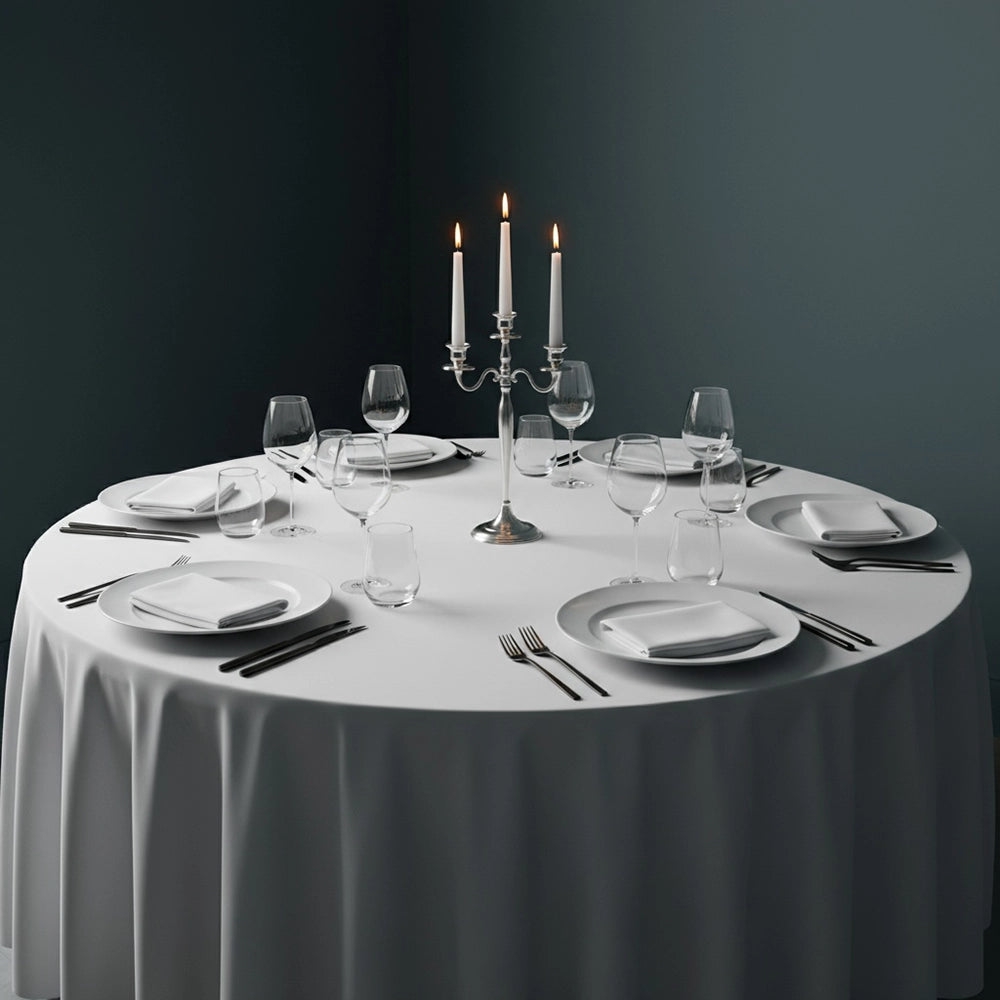 Elegant white round tablecloth on formal dining table set with white napkins, place settings, black cutlery, wine glasses, and silver candelabra against moody dark backdrop—luxury table linens by Linen & Co for hospitality and events